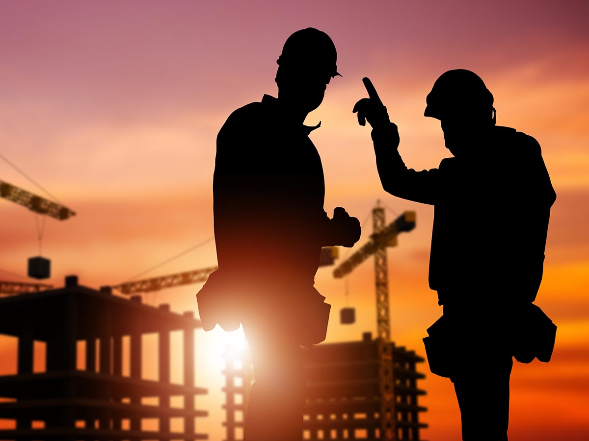 Two construction workers talking in the foreground, with the sunset casting colors over a building in construction.