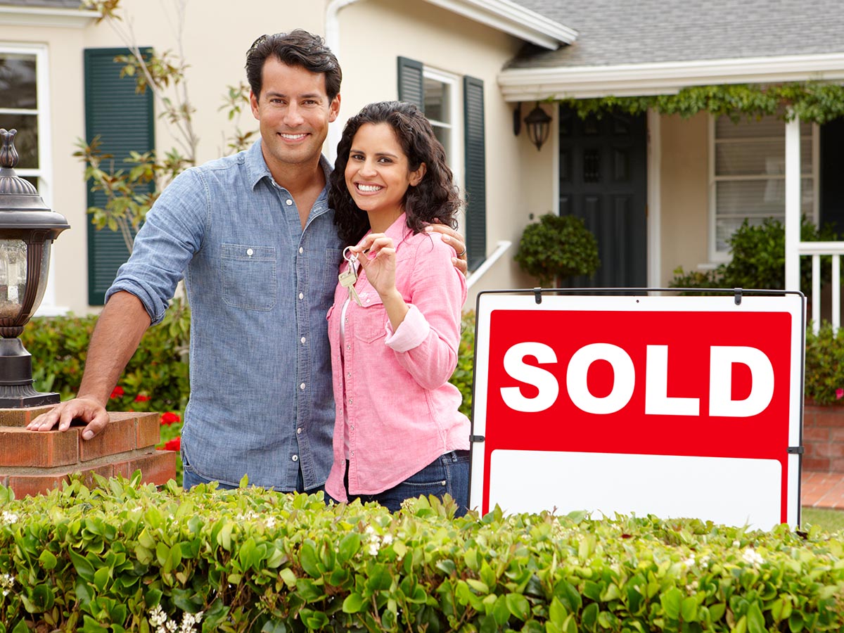 Happy couple standing in front of a house, with a “sold” sticker over a “For Sale” sign, holding the keys to the house, indicating they have just bought it. 